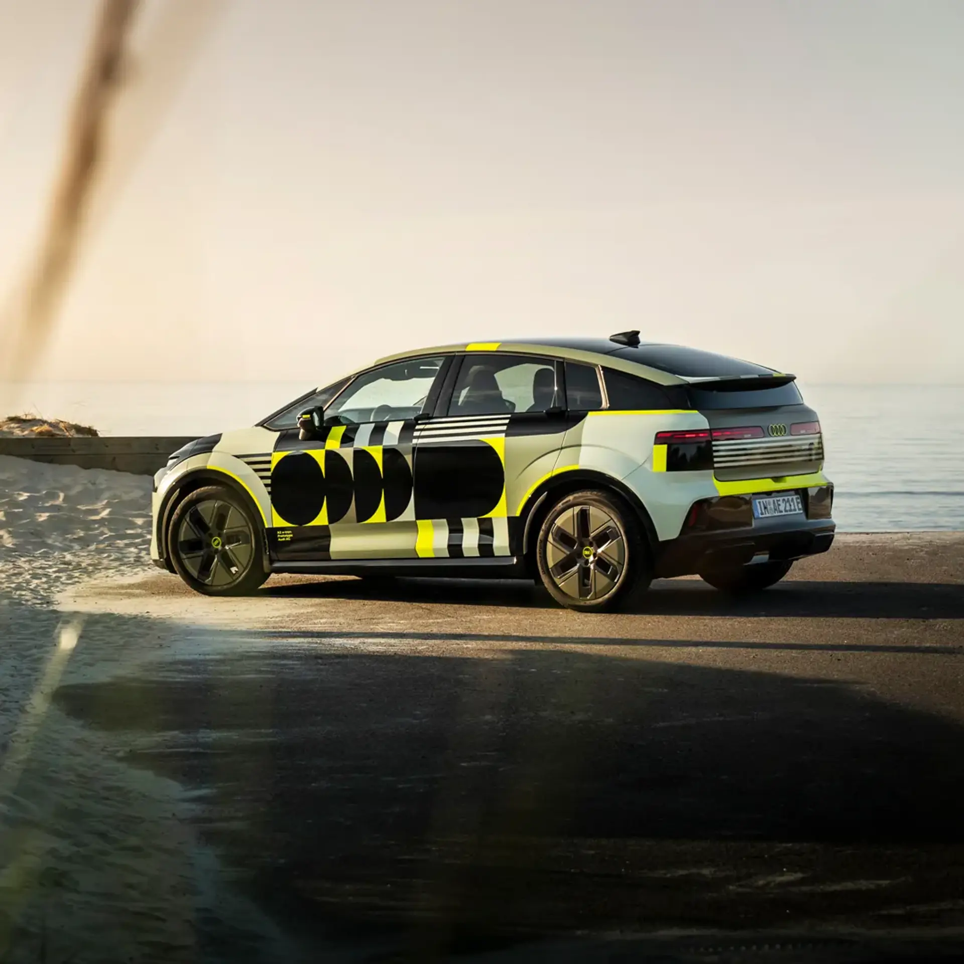 En moderne elbil med sort-gule grafiske striber holder parkeret ved en strand med havet i baggrunden.