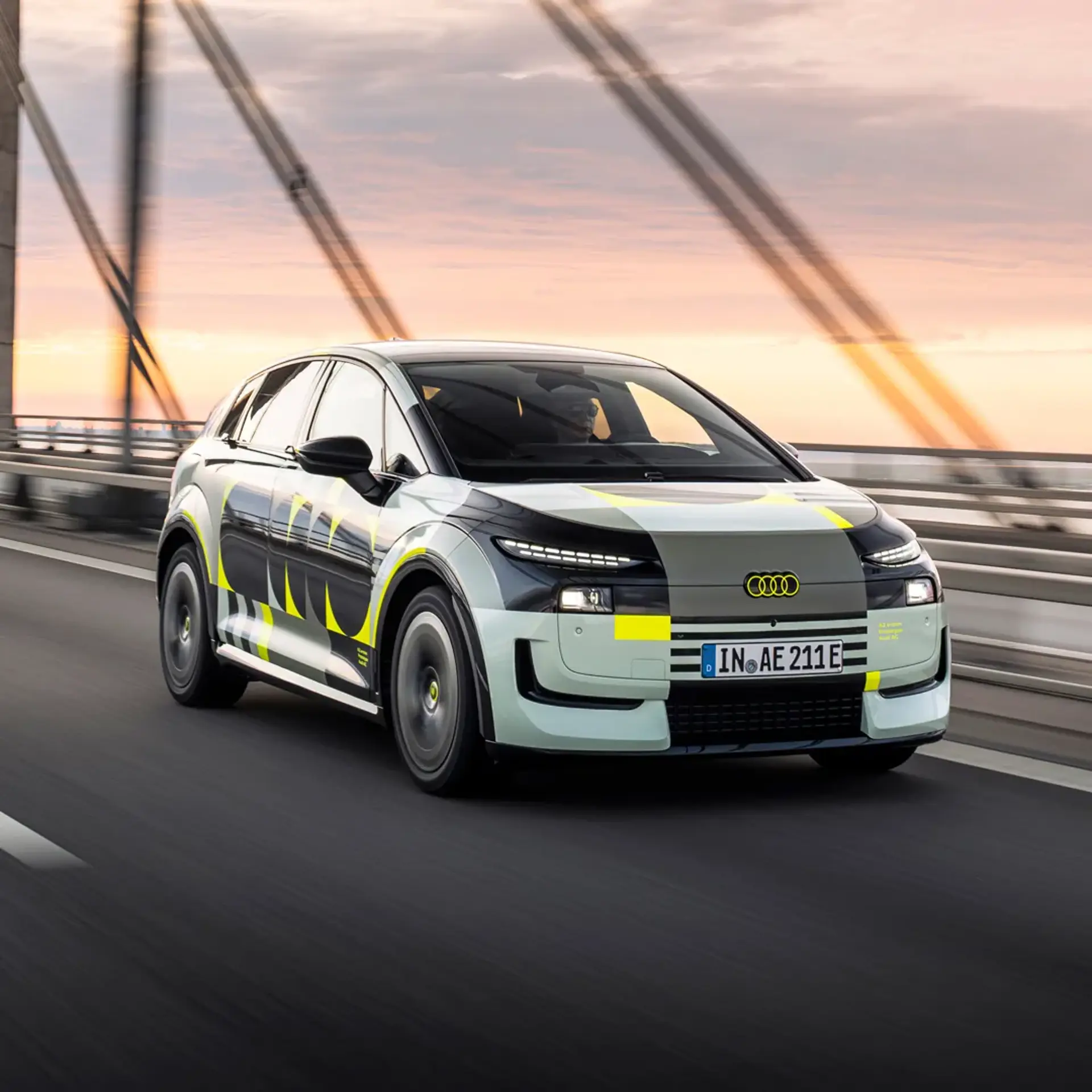 En moderne Audi elbil med grå og gule testmarkeringer kører over en bro i solnedgang.