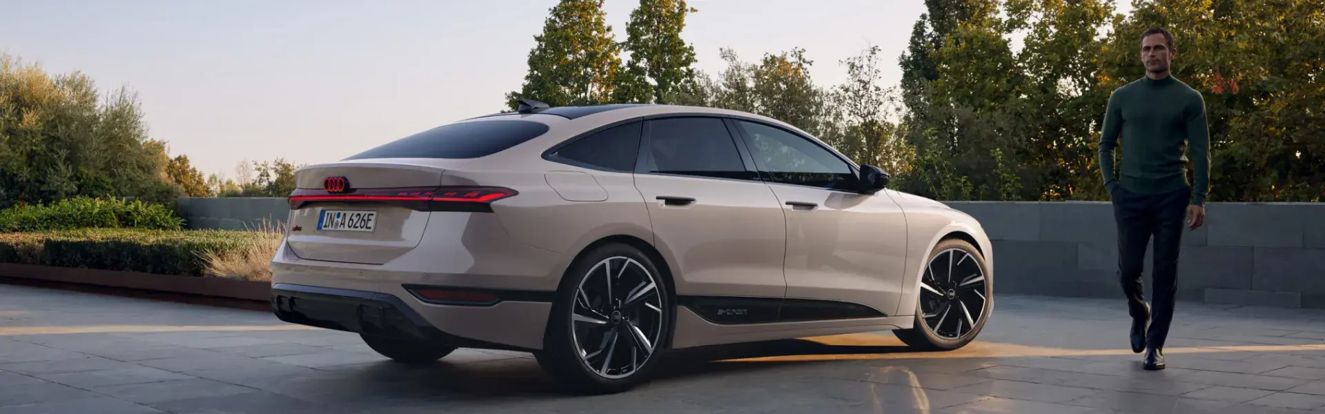 En lys Audi A6 Sportback e-tron holder parkeret udendørs ved en terrasse, mens en mand går forbi i baggrunden.