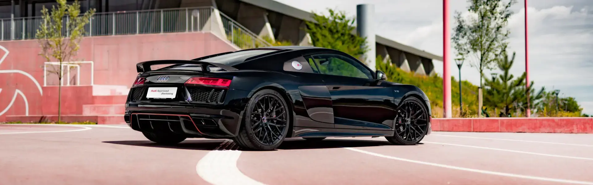 Sort Audi sportsvogn parkeret på en moderne, rød betonparkeringsplads med grønne træer i baggrunden.