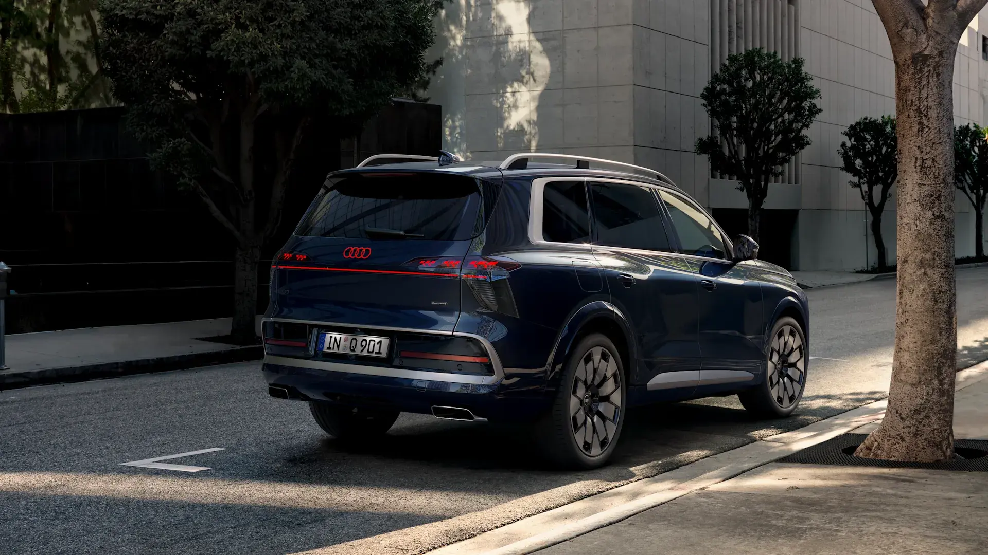 Blå Audi SUV set bagfra parkeret ved kantstenen i en by omgivet af træer og moderne bygninger.