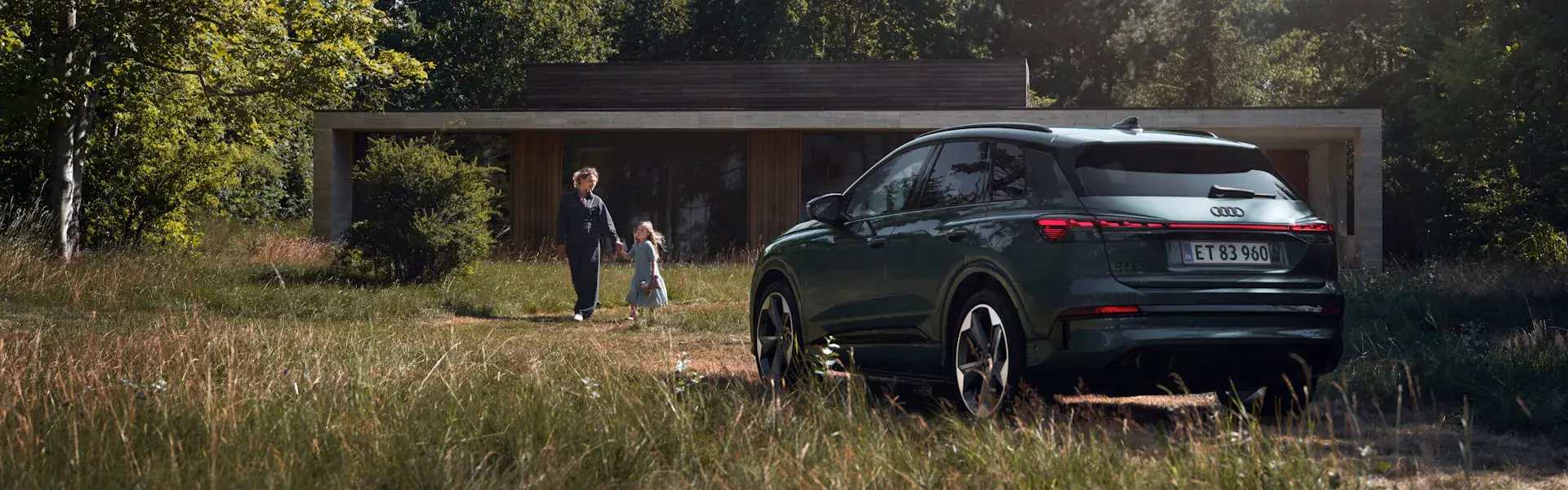 En mørkegrøn Audi SUV holder foran et moderne hus i naturen, mens en voksen og et barn går gennem det høje græs mod indgangen.