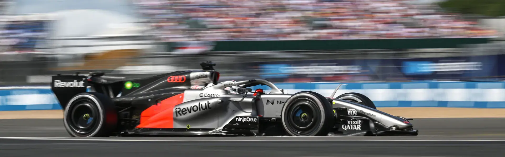 En Formel 1-racer i sølv og sort med sponsorlogoer kører i høj fart på en racerbane foran en sløret tilskuertribune.