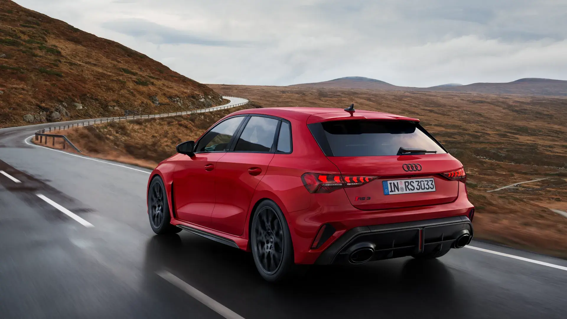 Rød Audi RS 3 Sportback ses bagfra i fart på en snoet bjergvej under overskyet himmel.