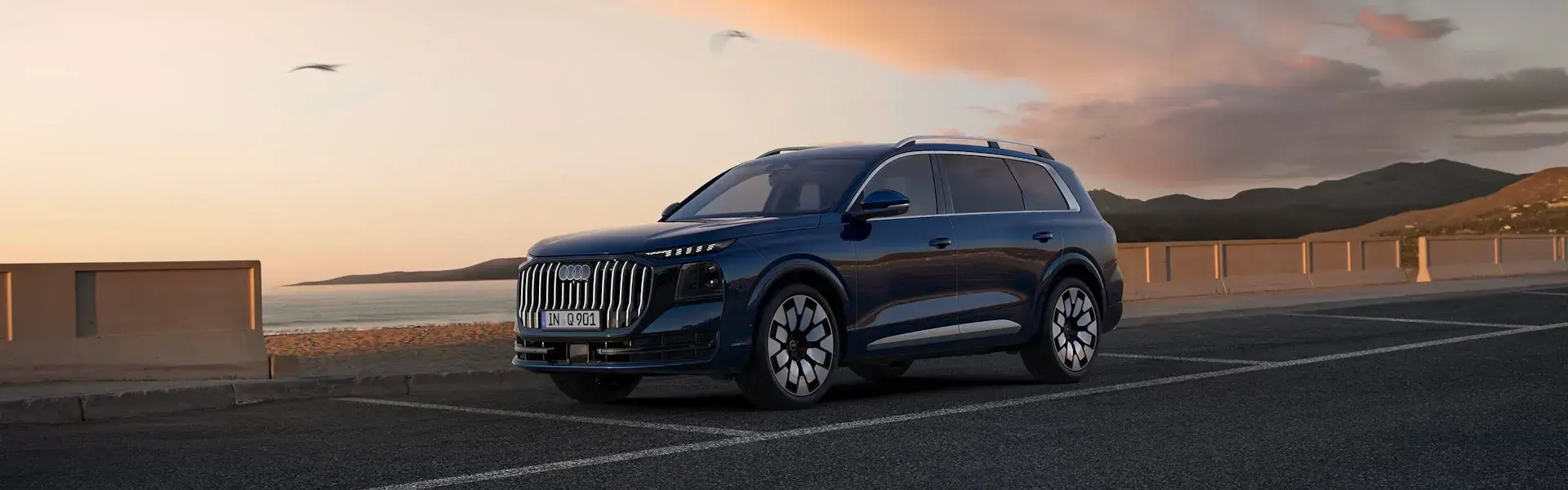 Mørkeblå SUV parkeret ved en kystvej i solnedgang med hav og bjerge i baggrunden.