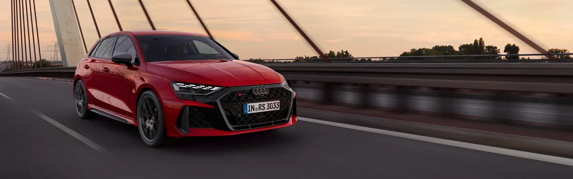Rød Audi RS 3 Sportback, der kører over en bro i aftensolen.