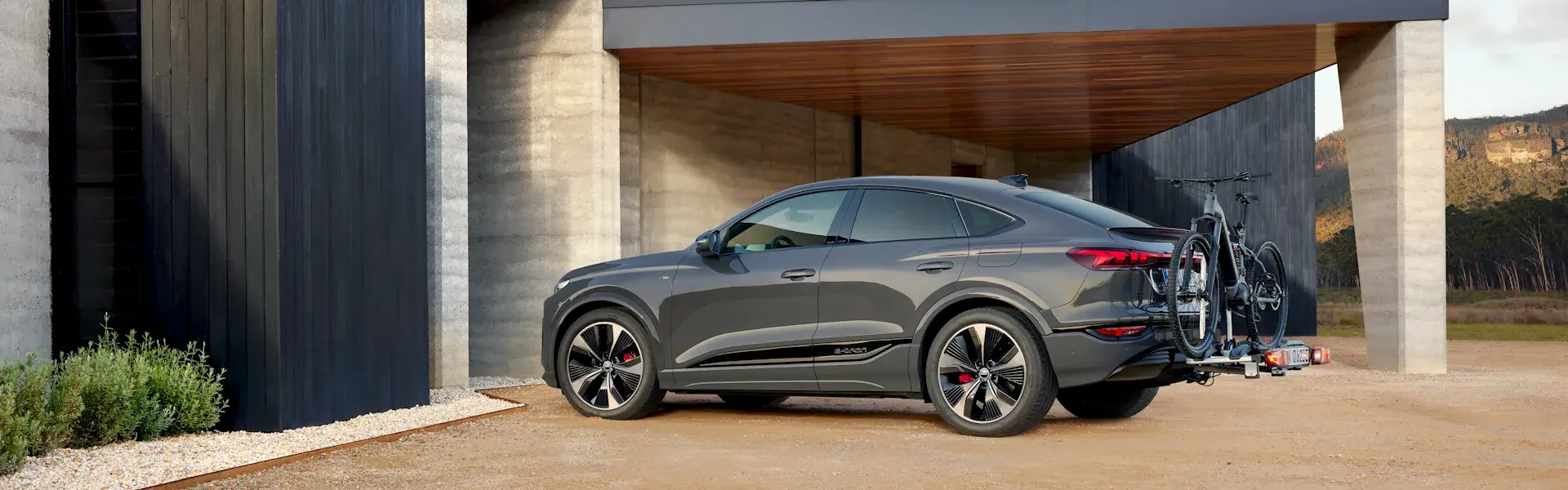 Grå Audi Q6 e-tron med cykelholder og to cykler monteret bagpå holder parkeret ved et moderne hus i naturen.